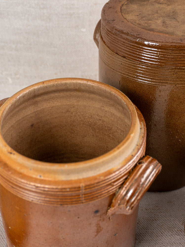 Three antique French stoneware confit pots à graisse, some lidded and handled, on a textured beige surface.