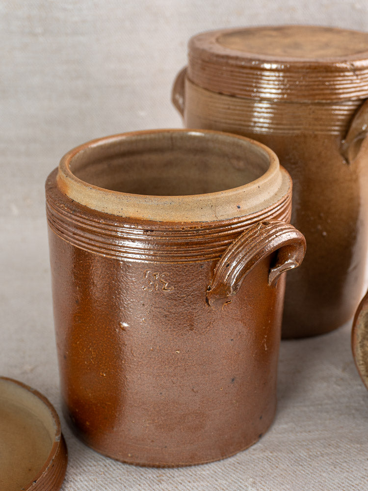 Set of three antique French stoneware confit pots with lids, rustic textured finish, one pot open in front.