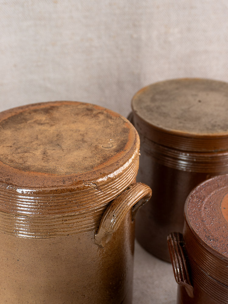 Three antique French stoneware confit pots with textured lids and handles on a neutral fabric surface.