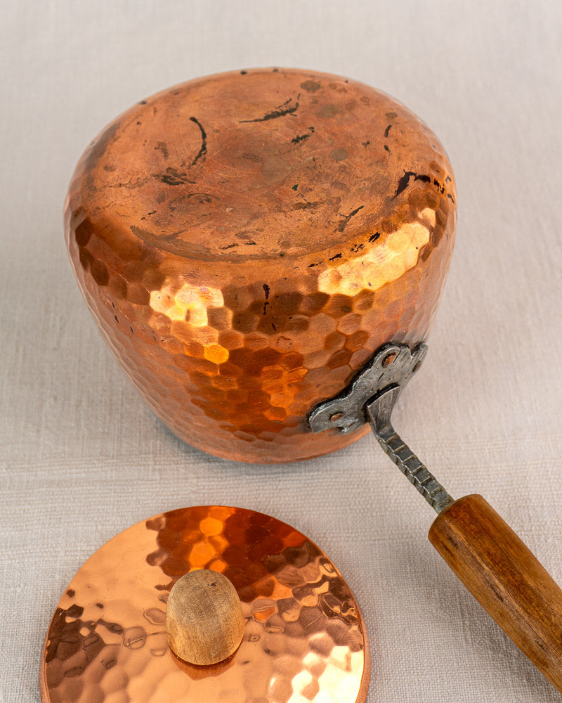 Antique French hammered copper fondue set with wooden handle, copper lid, and iron stand on a light textured surface.