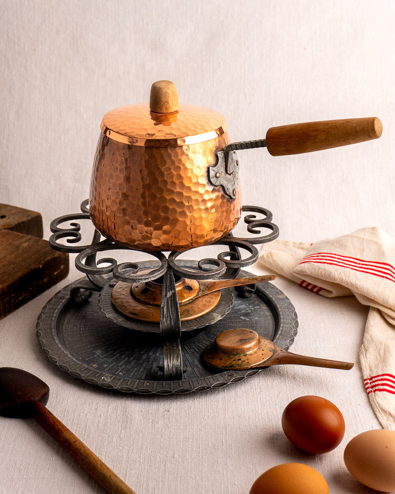 Antique French copper fondue set with hammered pot and iron stand, on neutral tablecloth with wooden spoon and eggs.