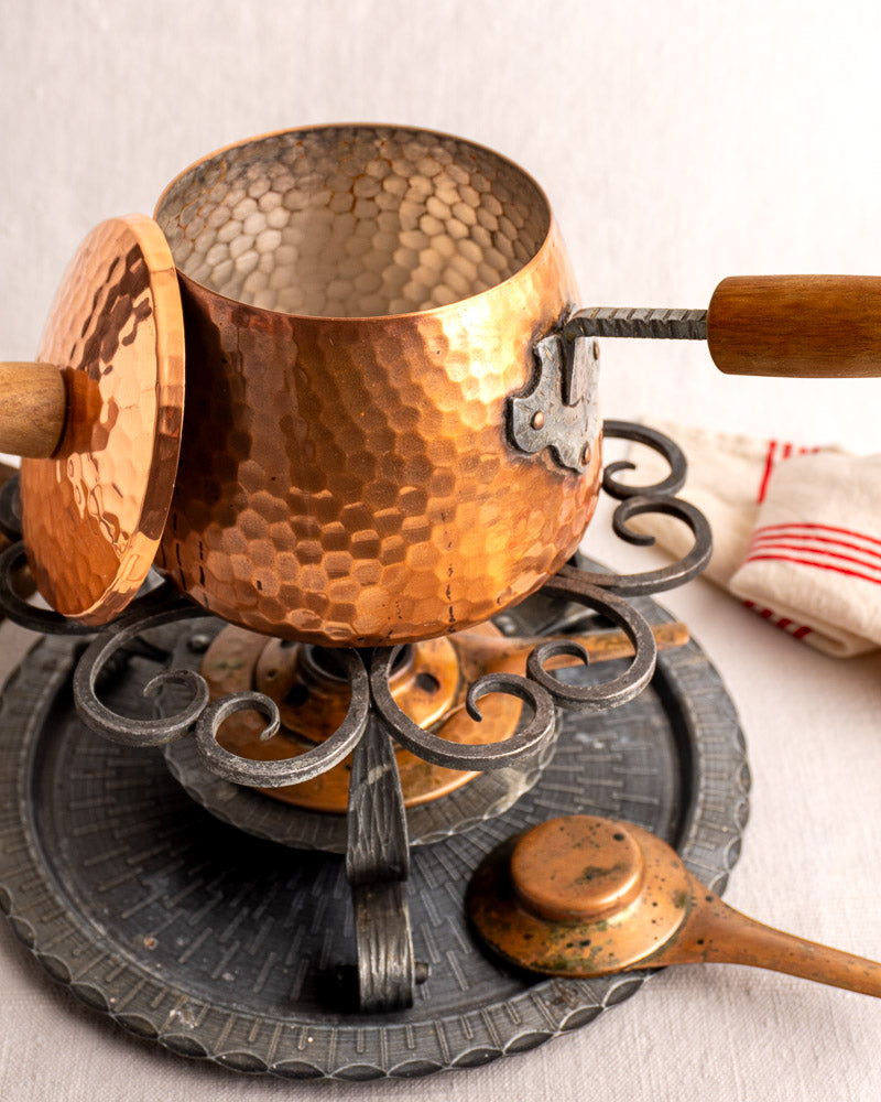 Antique French copper fondue set with hammered pot, wood handle, iron stand, and tray, red-striped cloth background.