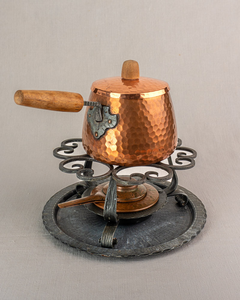 Antique French copper fondue set with hammered pot, wood handle, ornate iron stand, and round metal tray on plain background.
