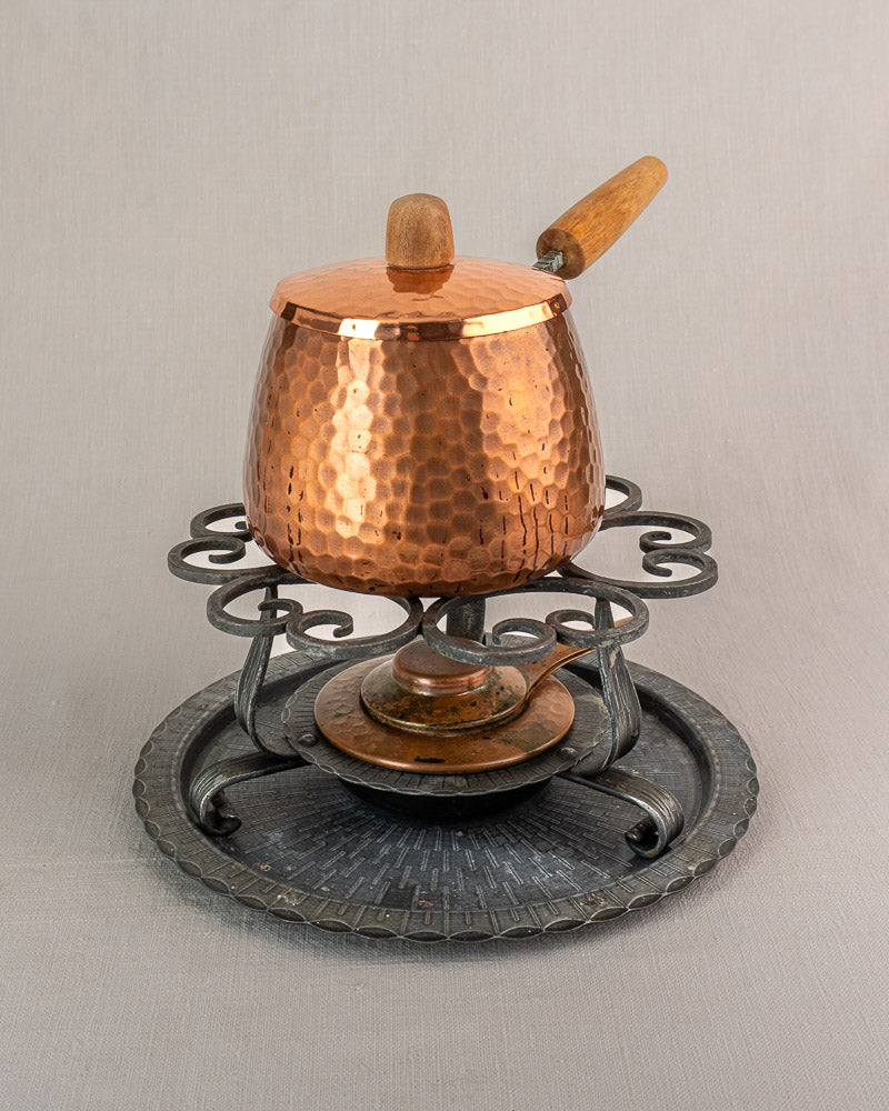 Antique French copper fondue set with hammered pot and wooden handle on ornate iron warmer, light gray background.
