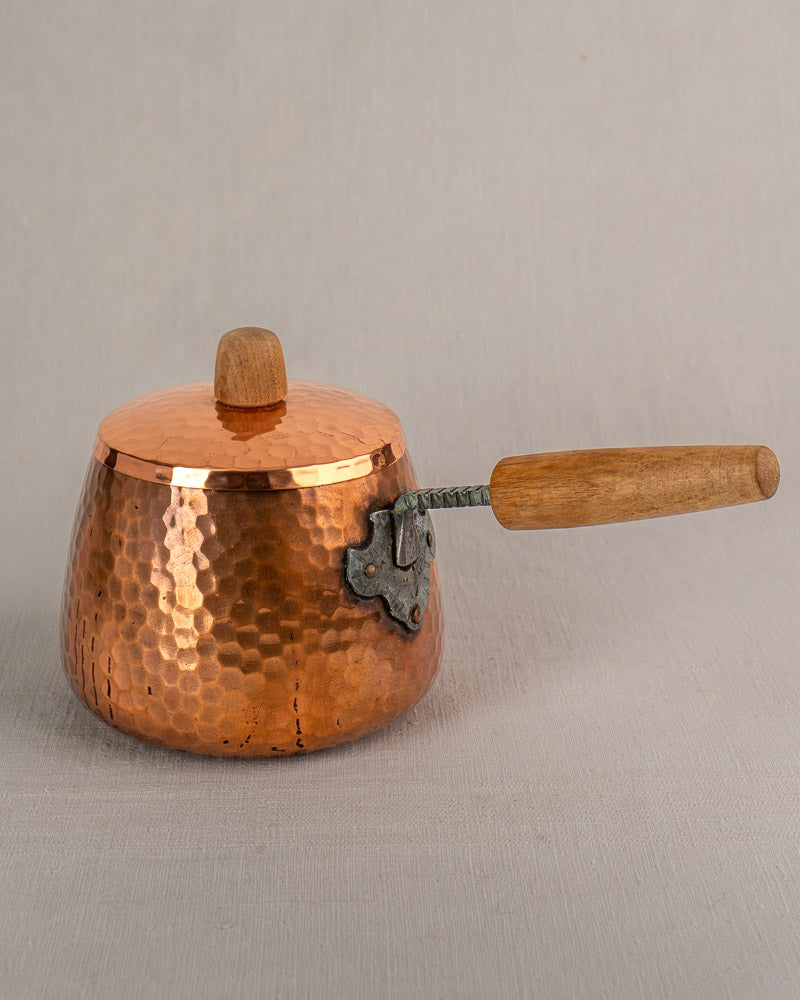 Antique French copper fondue set with hammered pot, lid, and wooden handle on iron stand against neutral gray surface.