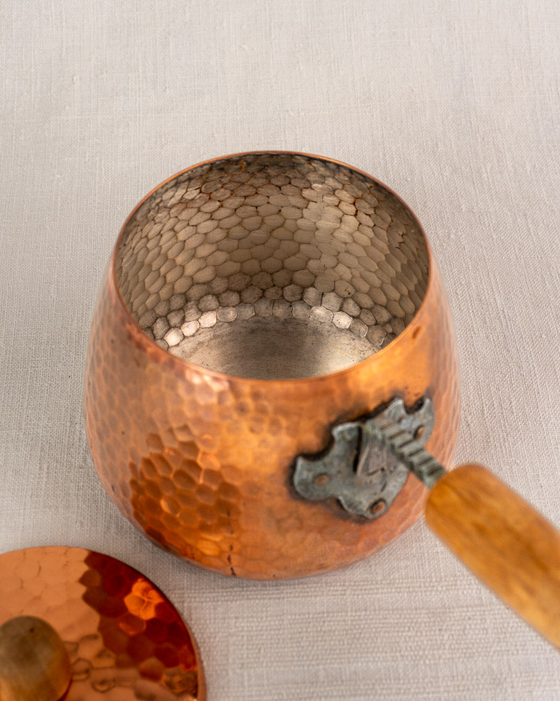 Antique French hammered copper fondue set with metal handle, lid, and iron warmer on light fabric.