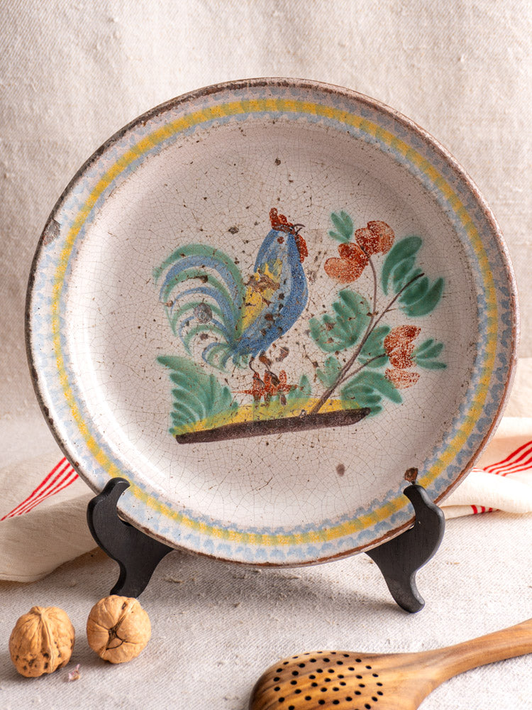 Antique French faience plate with hand-painted blue and red rooster, on black stand with beige cloth, wooden spoon, and walnuts.