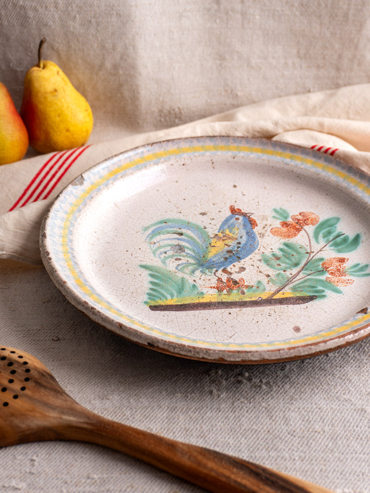 Antique French faience plate with hand-painted rooster, displayed on textured cloth beside wooden spoon and yellow pears.