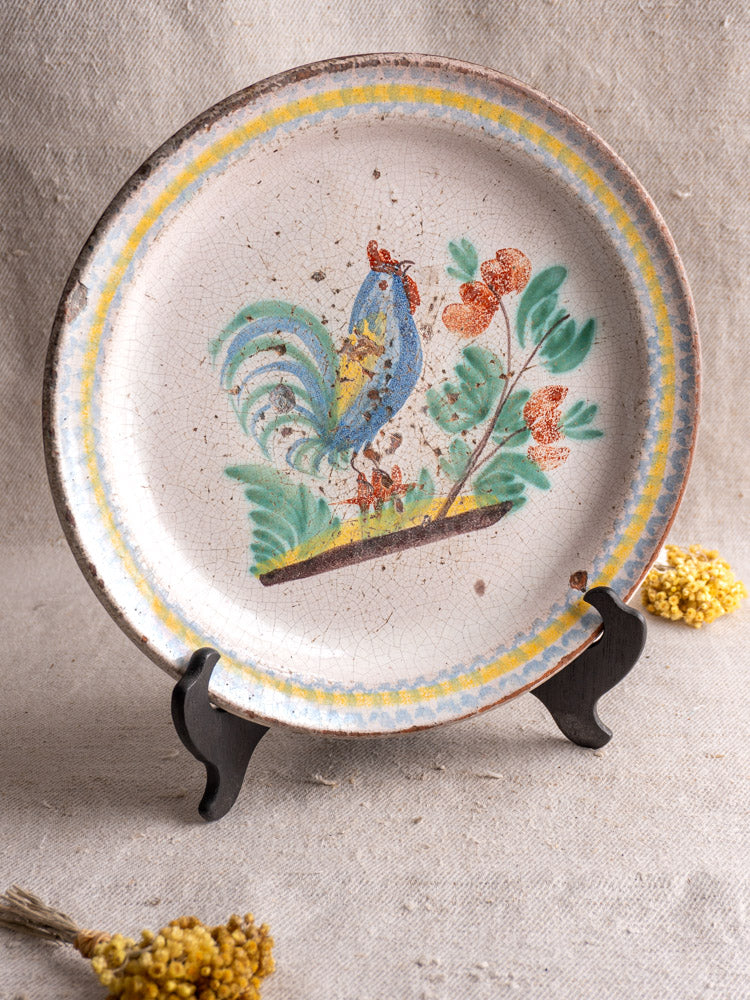 Antique French faience plate with hand-painted rooster and branch, displayed upright near dried yellow flowers. Late 19th century.