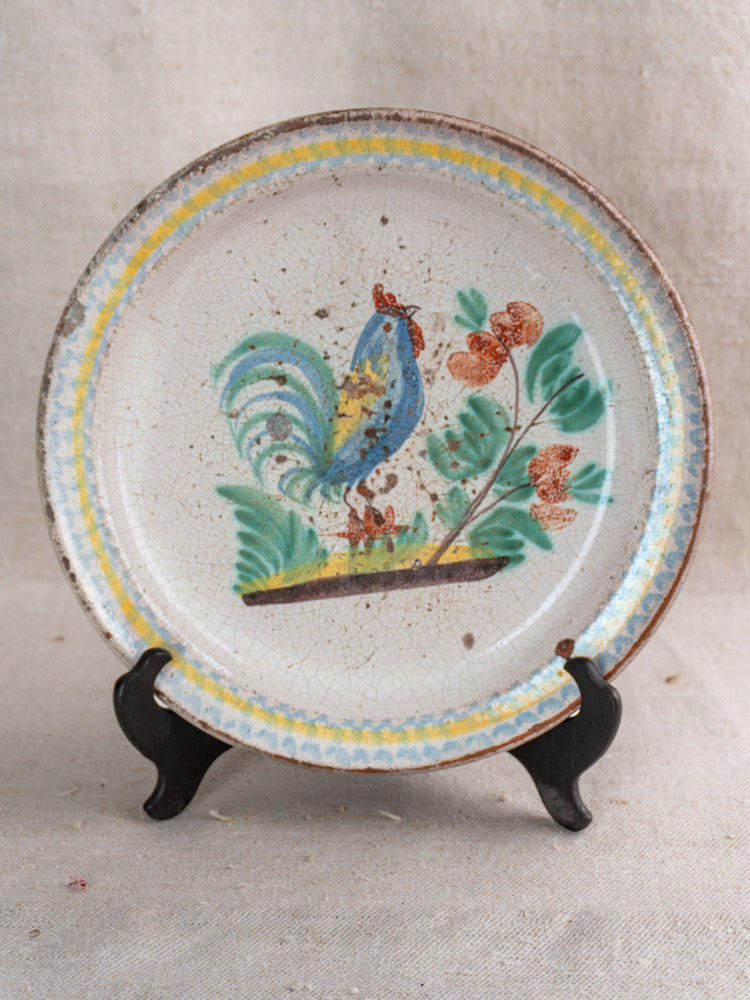 Late 19th-century French faience plate, hand-painted with a blue and yellow rooster, displayed on black stand.