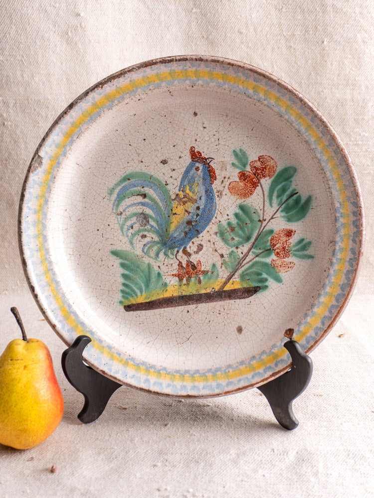 Antique French faience plate with hand-painted rooster, displayed on black stand beside a pear, light fabric backdrop.