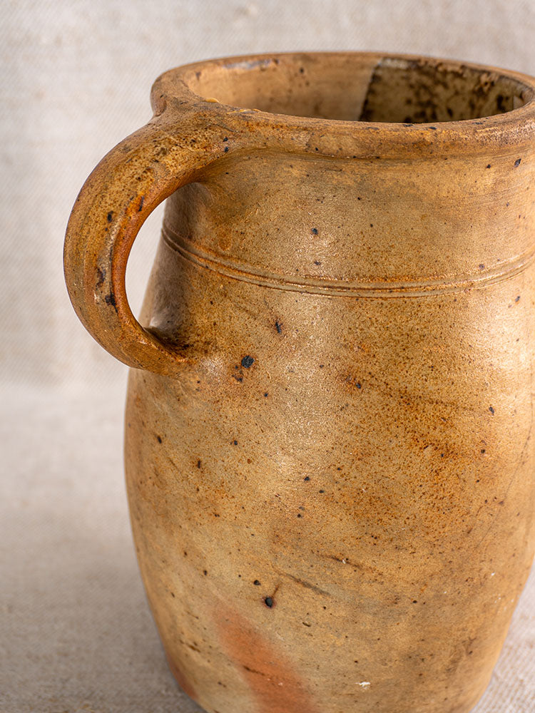 Early 19th-century French stoneware pitcher with single handle, salt-glazed finish, on neutral woven fabric background.