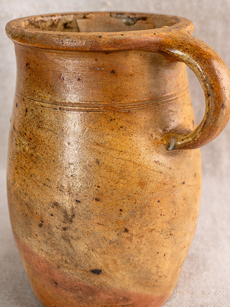 Antique French stoneware pitcher, early 19th century, with handle and weathered salt glaze on neutral fabric background.