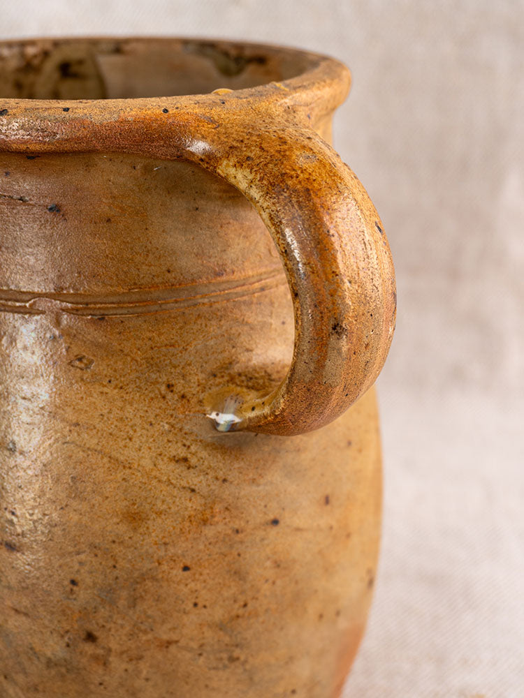 Antique French salt glazed stoneware pitcher with rustic handle and minor base chip on textured neutral fabric background.
