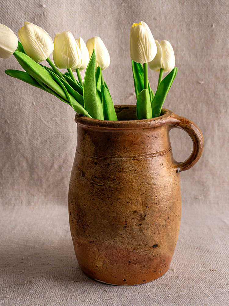 Antique French salt-glazed stoneware pitcher with handle, filled with white tulips, on a neutral textured fabric background.