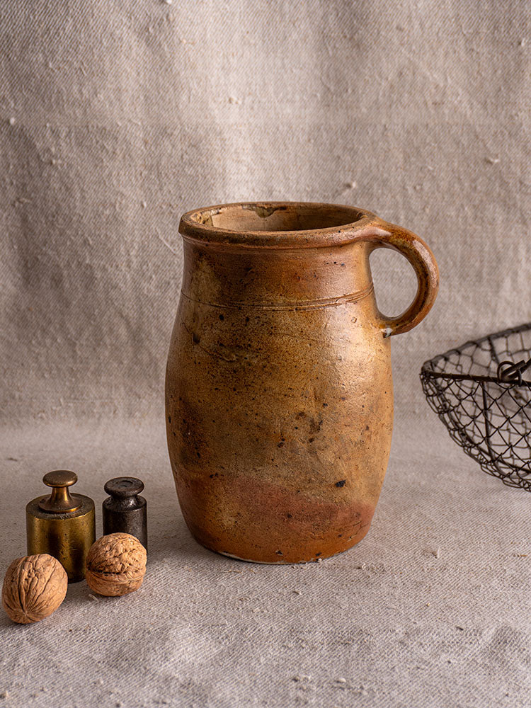 Antique French salt glazed stoneware pitcher with handle, on beige surface with walnuts, metal weights, and wire basket nearby.