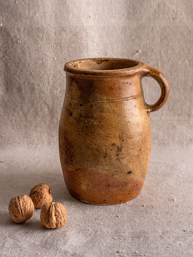 Antique French salt-glazed stoneware pitcher with handle on beige fabric, three whole walnuts beside, neutral backdrop.
