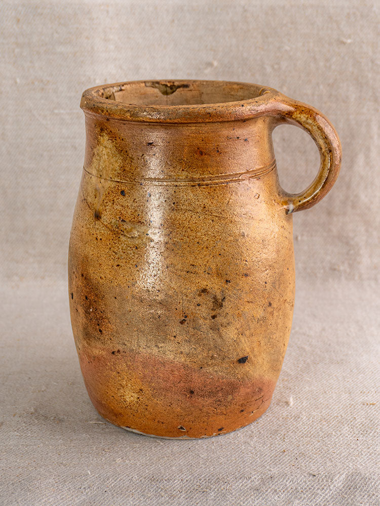 Antique French stoneware pitcher with single handle and rustic brown glaze on textured light fabric.
