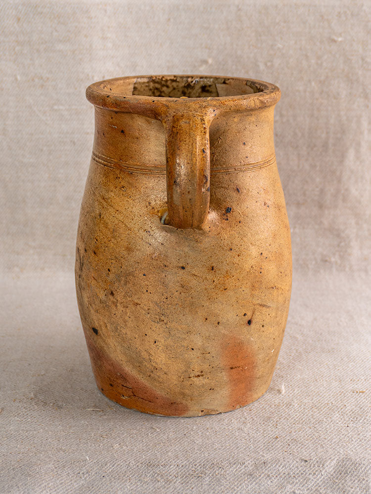 Antique French salt-glazed stoneware pitcher, rustic brown with rounded handle, displayed on textured beige fabric.