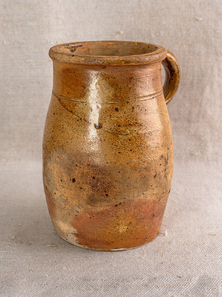 Antique French stoneware pitcher with salt-glazed finish and textured surface on beige fabric background.