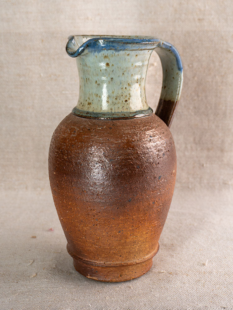 Antique French stoneware pitcher with textured brown base, blue ash celadon upper section, on beige fabric.