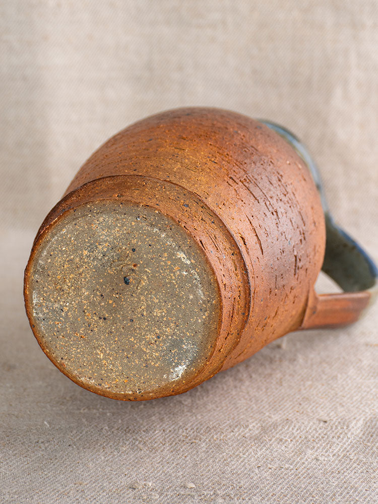 French antique stoneware pitcher from La Borne with blue ash celadon glaze, shown on neutral woven fabric.