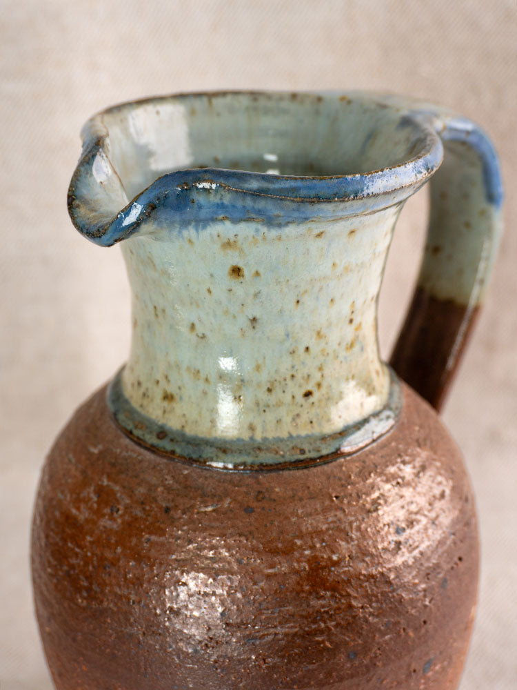 Antique French stoneware pitcher with wide spout, rounded handle, and blue ash celadon glaze on beige fabric background.