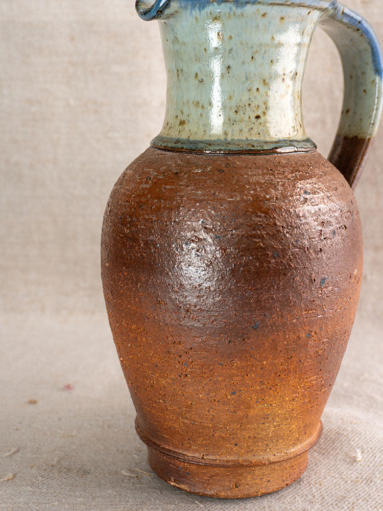 Antique French stoneware pitcher with blue ash celadon glazed top and handle on neutral fabric background.