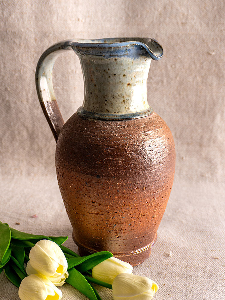 Antique French stoneware pitcher with blue ash celadon glaze, displayed on beige surface beside yellow tulips.
