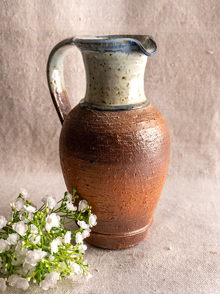 Antique French stoneware pitcher from La Borne with blue ash celadon glaze, on beige textured surface with white flowers.