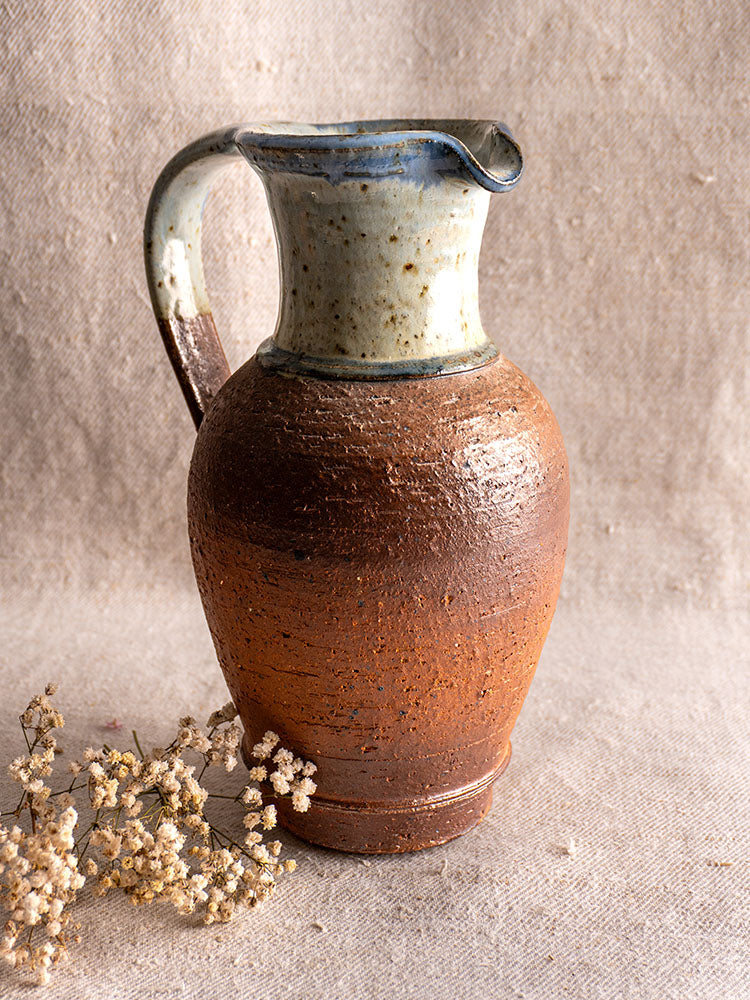Antique French La Borne stoneware pitcher with blue ash celadon glaze on beige fabric near dried white flowers.