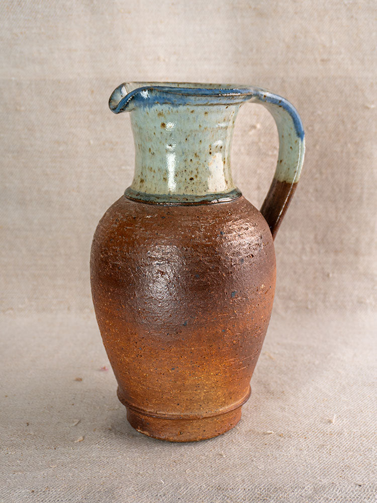 Antique French stoneware pitcher with rounded body, narrow neck, brown base, blue ash celadon glaze on fabric background.