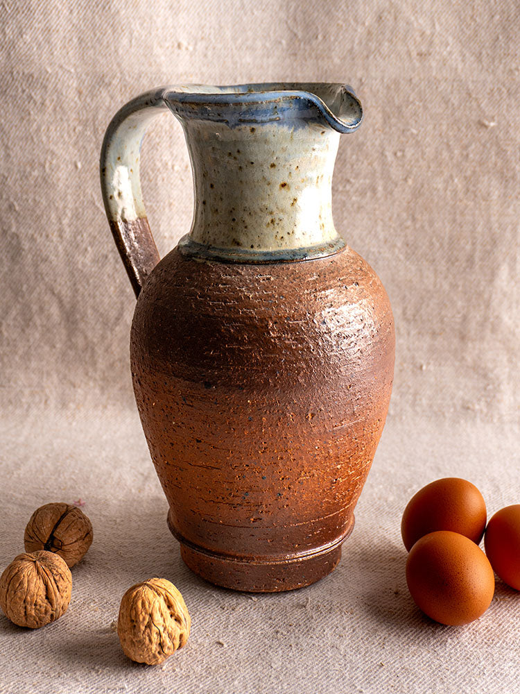 Antique French stoneware pitcher with rounded body, narrow neck, brown base, blue ash celadon glaze on fabric background.