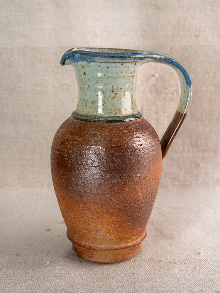 Antique French stoneware pitcher with rustic brown body and blue ash celadon rim, on neutral fabric background.