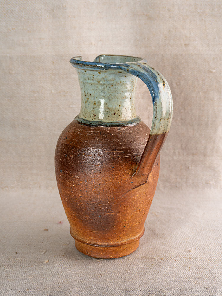 Antique French La Borne stoneware pitcher with brown body, blue ash celadon neck, on textured beige fabric.