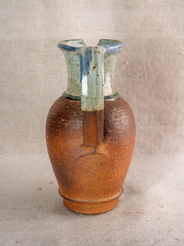 Antique French La Borne stoneware pitcher with narrow neck, blue celadon glaze, on textured beige fabric.
