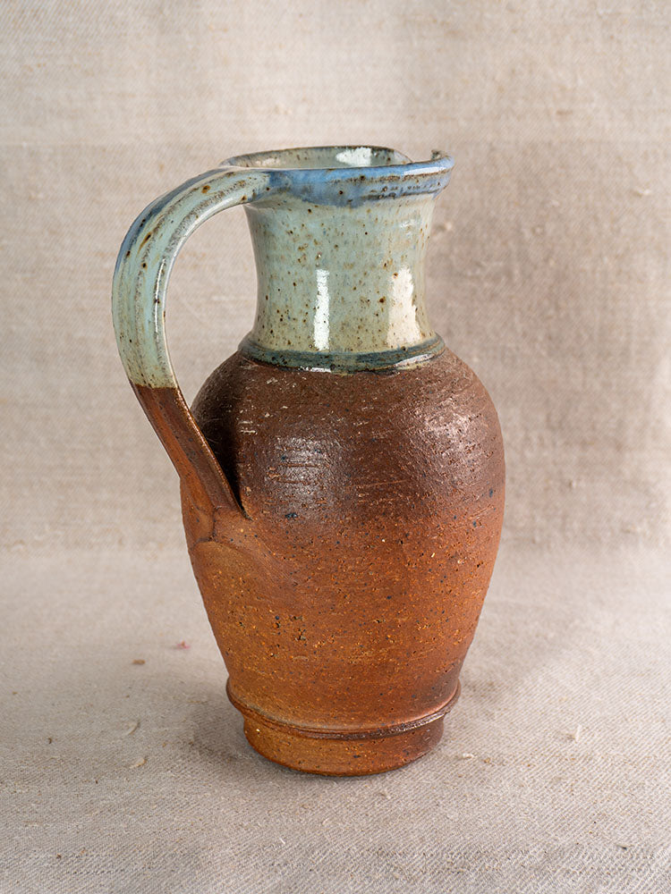 Antique French stoneware pitcher with blue ash celadon glaze and textured brown base, on neutral fabric background.