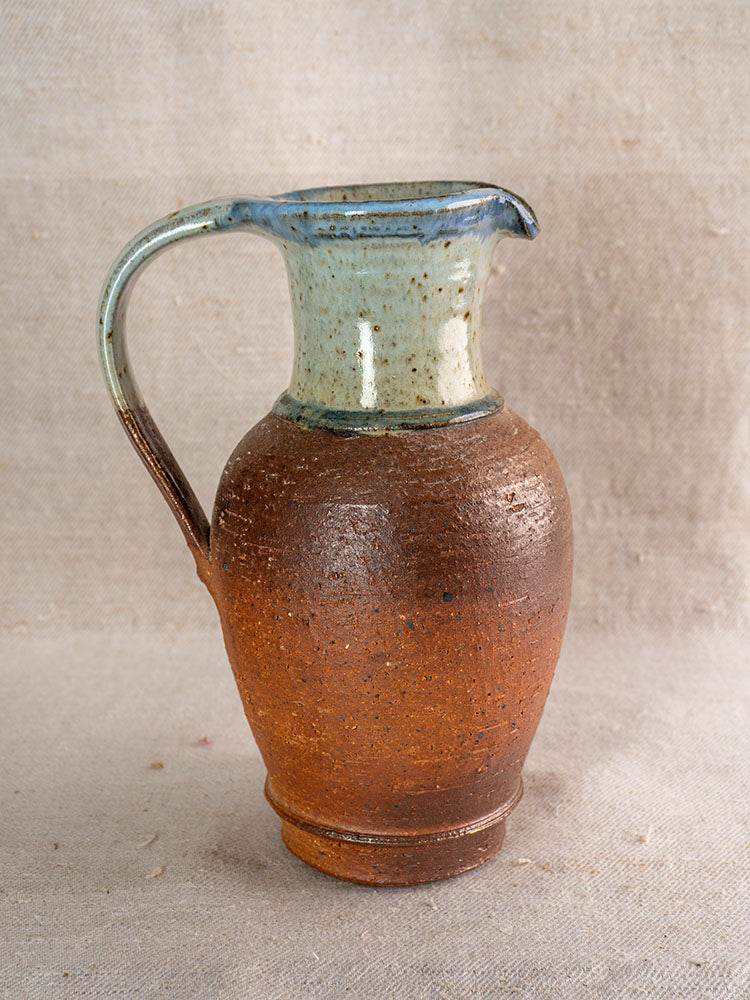 Antique French stoneware pitcher with mottled brown base, blue ash celadon glaze on textured neutral fabric background.