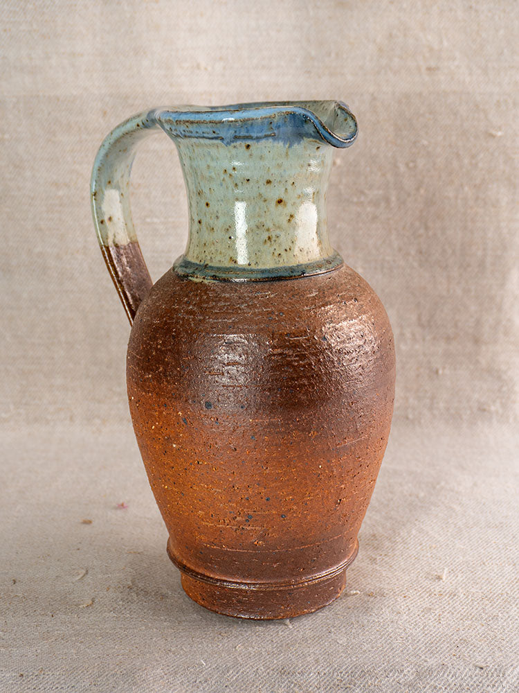 French stoneware pitcher with blue celadon glazed neck, rounded brown base on textured neutral fabric background.