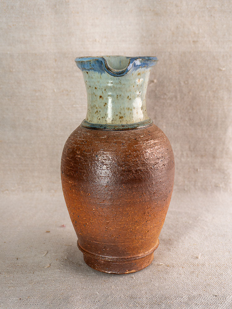 Antique French stoneware pitcher with brown textured body, blue ash celadon neck, on neutral fabric background.