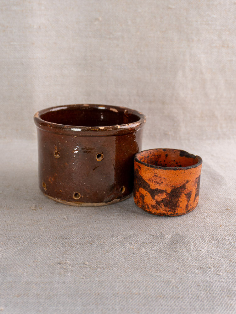 Pair of 19th century French ceramic cheese strainers, brown and orange, shown on textured beige fabric.