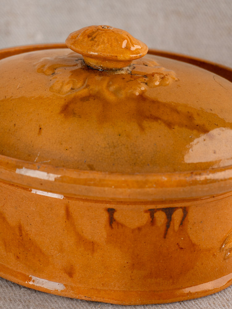 Antique French terrine with mustard ochre glaze, round ceramic body and lid, on neutral fabric background.