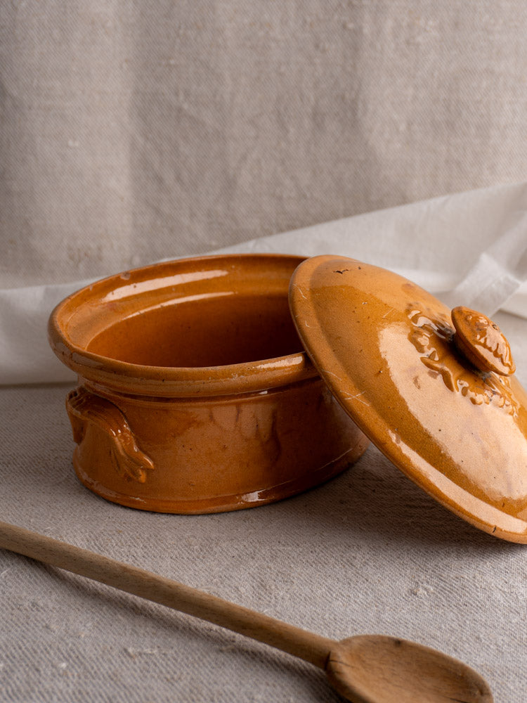 Antique French terrine with mustard ochre glaze, late 19th–early 20th century, on beige cloth beside wooden spoon.