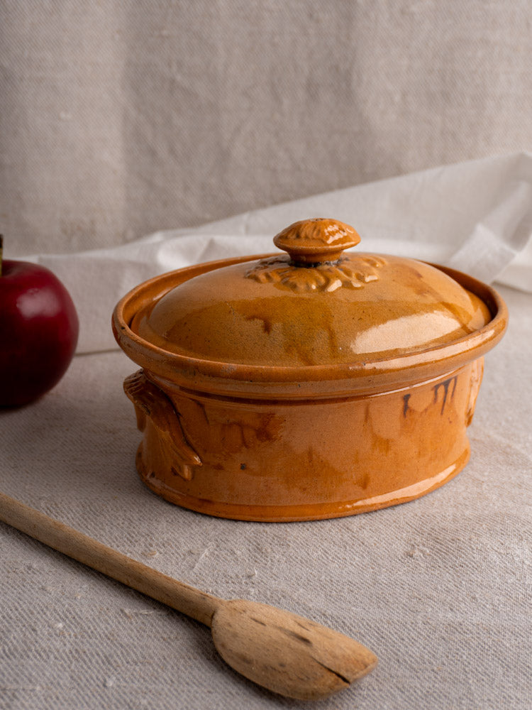 Antique French terrine with mustard ochre glaze on beige cloth beside wooden spoon and red apple.