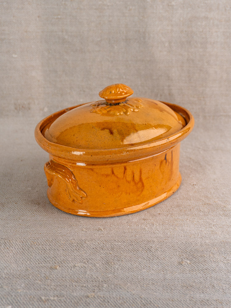 Antique French terrine with mustard ochre glaze, oval shape, decorative handles, on beige textured fabric background.