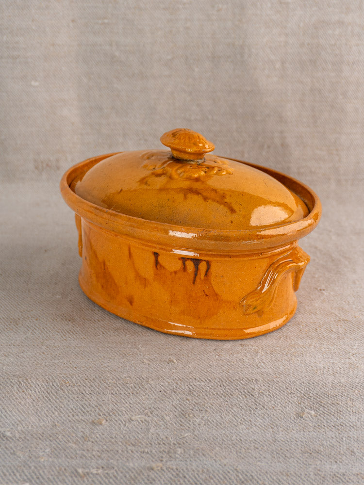 Antique French terrine with mustard ochre glaze, oval shape, decorative handles, on textured beige fabric background.
