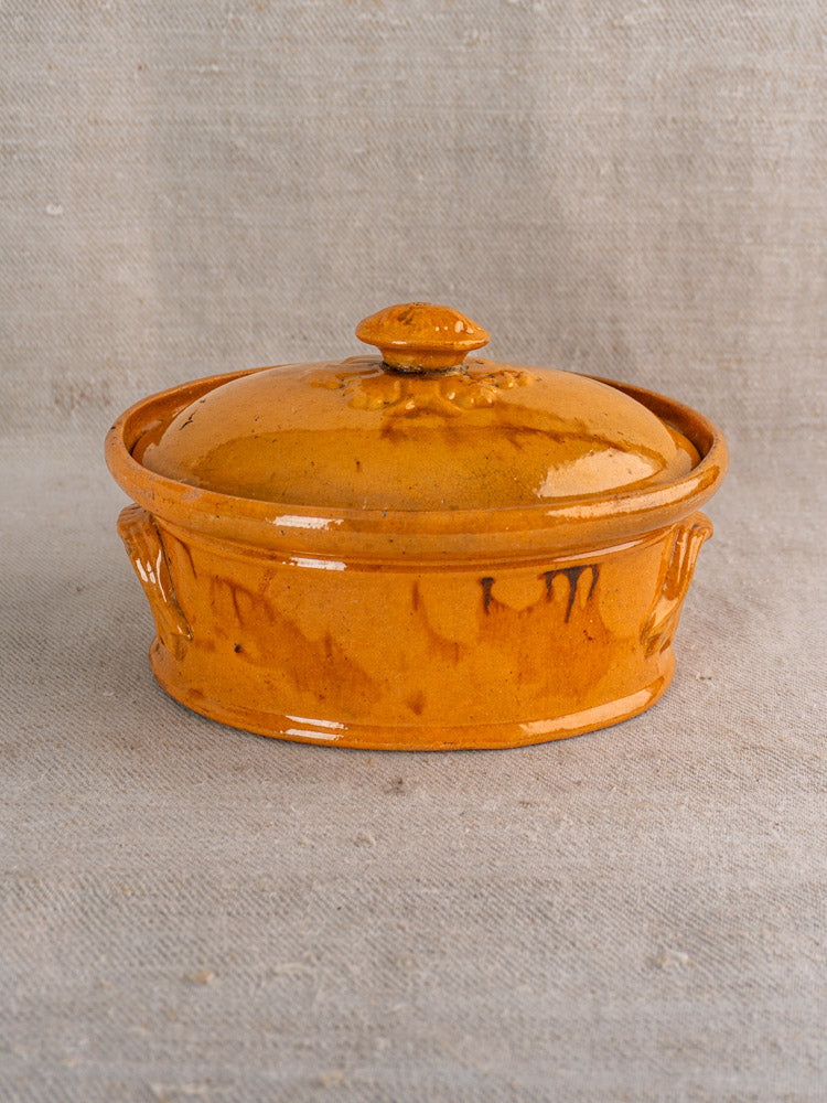 Antique French terrine with mustard ochre glaze, side handles, and lid on textured beige fabric. Late 19th–early 20th century.