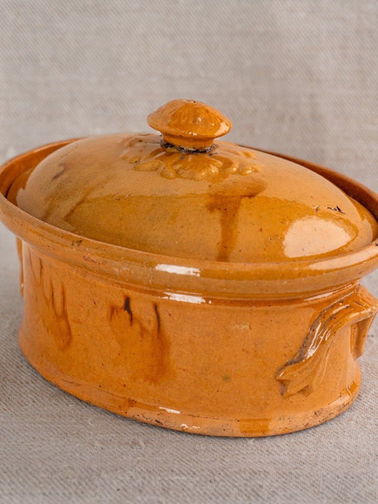 Antique French oval brown ceramic terrine with mustard ochre glaze and decorative handle on textured gray fabric.