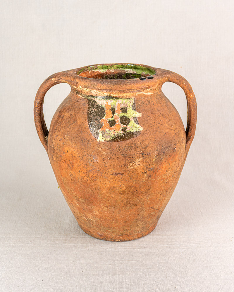 Antique French glazed terracotta vinaigrier with two handles, rounded form, and aged patina on fabric background.