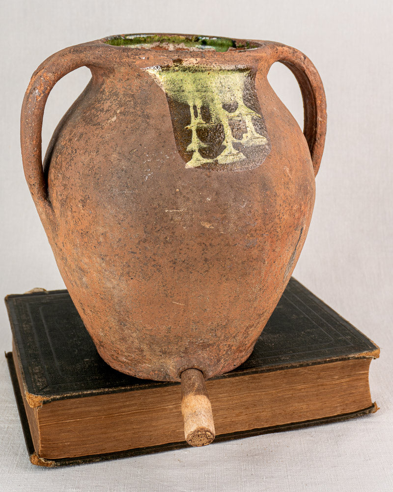 Antique French glazed terracotta vinaigrier with two handles, rounded form, and aged patina on fabric background.
